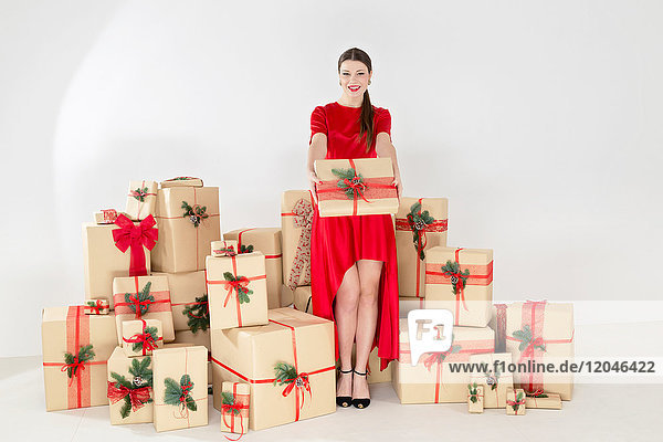 Porträt einer jungen Frau in rotem Kleid durch gestapelte Weihnachtsgeschenke beim Überreichen eines Weihnachtspakets