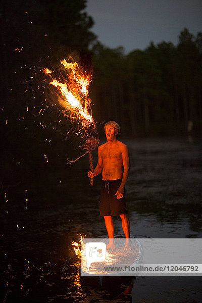 Junger Mann steht in der Dämmerung auf einem Paddelbrett auf dem Wasser  hält brennenden Ast  Flammen auf dem Paddelbrett