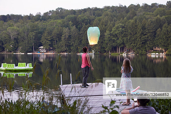 Girl standing on jetty releasing sky lantern  young girl watching  woman photographing event using smartphone