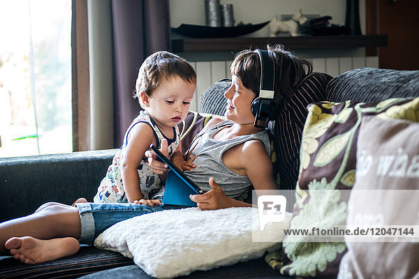 Neugieriges männliches Kleinkind betrachtet das digitale Tablet des Bruders auf dem Sofa