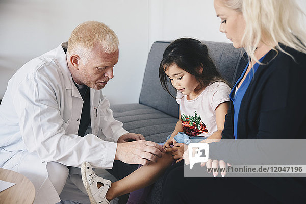Älterer Arzt  der das Knie des Mädchens untersucht  während die Mutter im Krankenhaus auf dem Sofa sitzt.