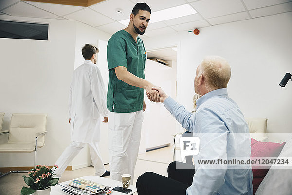 Young male nurse shaking hands with mature patient sitting in lobby at hospital
