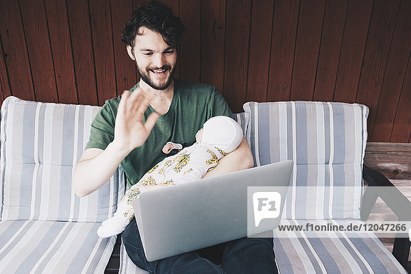 Lächelnder Vater mit Babyjunge bei einer Videokonferenz am Laptop in der Ferienvilla