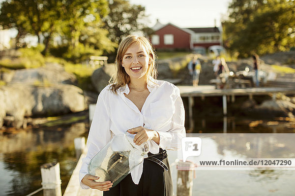 Porträt einer lächelnden jungen Frau,  die ein Glas am Steg des Hafens hält.