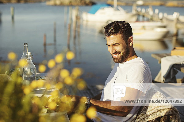 Lächelnder junger Mann sitzt auf Stuhl am Tisch gegen den See