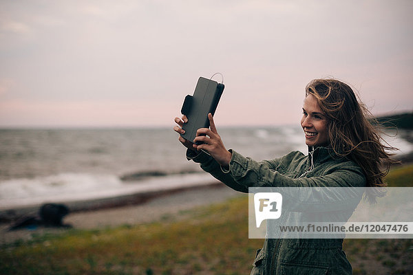 Lächelnde junge Frau nimmt Selfie auf digitalem Tablett am Strand mit.