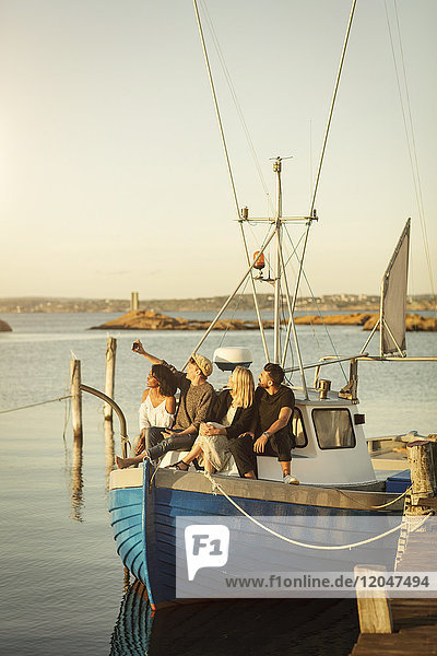 Junger Mann nimmt Selfie mit Freunden mit  während er auf einem Boot sitzt  das am Hafen festgemacht ist.