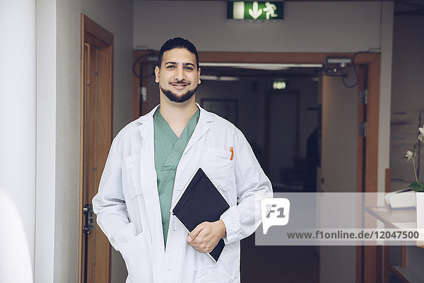 Portrait of smiling young male doctor holding notepad while standing in hospital corridor
