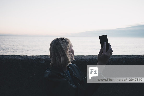 Frau nimmt Selfie durch Handy  während sie bei Sonnenuntergang am See steht.
