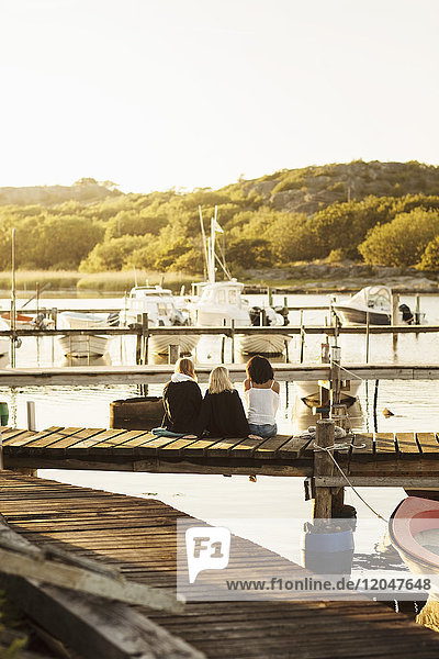 Rückansicht von jungen Freundinnen  die den Sommer genießen  während sie am Steg am Hafen gegen den Himmel sitzen.