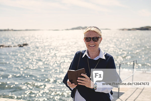 Porträt einer lächelnden Seniorin mit Sonnenbrille  die ihr Handy gegen den See hält.