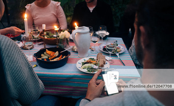 Beschnittenes Bild eines jungen Mannes  der ein Smartphone benutzt  während er mit Freunden zu Abend isst.