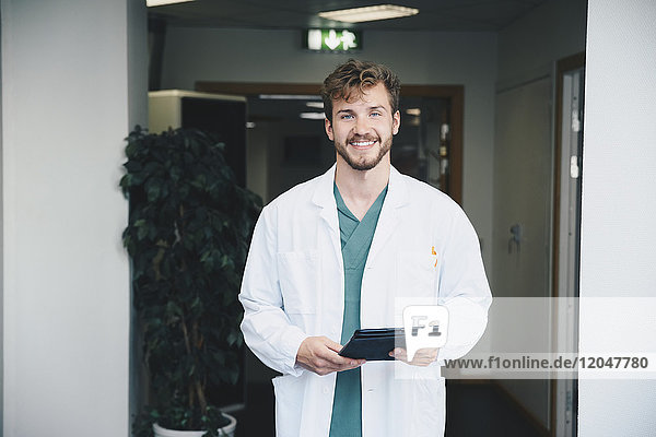 Portrait of confident smiling male doctor standing with digital tablet at hospital corridor