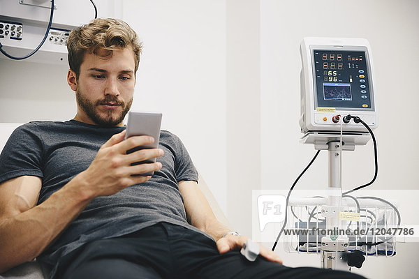 Young male patient using smart phone while reclining on bed during medical test in hospital