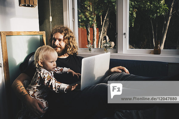 Vater mit Mädchen hält Laptop beim Sitzen am Fenster zu Hause