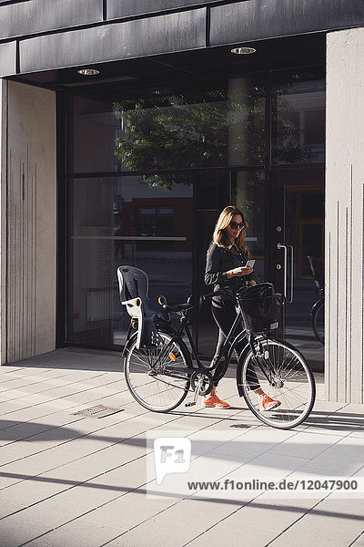 Reife Frau  die mit dem Fahrrad unterwegs ist  während sie das Smartphone auf dem Bürgersteig gegen das Gebäude in der Stadt benutzt.