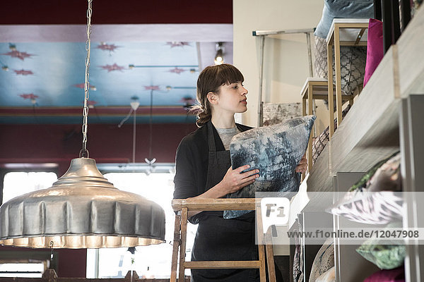 Low angle view of young female entrepreneur standing on ladder while arranging cushion in rack at store