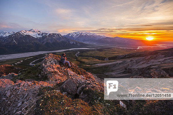 Ein Mann beobachtet einen ruhigen Sonnenuntergang von einem alpinen Sitzplatz hoch über dem Delta River in der Alaska Range; Alaska  Vereinigte Staaten von Amerika
