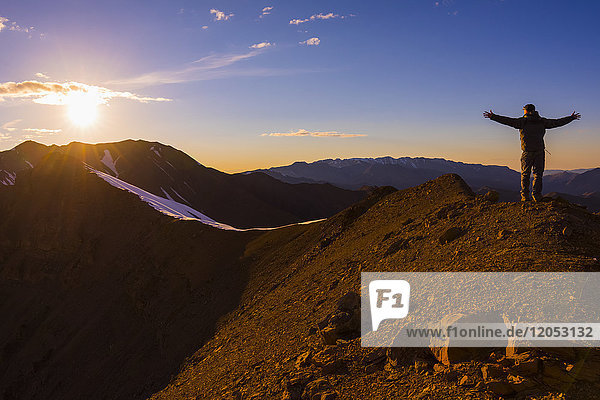 Ein Mann steht mit ausgestreckten Armen auf einem Bergrücken  während die Sonne über dem Polychrome Mountain im Denali National Park untergeht; Alaska  Vereinigte Staaten von Amerika