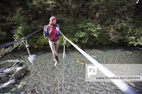 Eine Frau geht auf einem Kabel auf einer Hängebrücke über einen Fluss; Alaska  Vereinigte Staaten von Amerika
