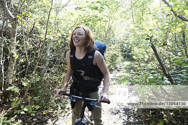 Eine Frau steht auf einem Pfad in einem Wald mit ihrem Fahrrad und einem Rucksack; Alaska  Vereinigte Staaten von Amerika