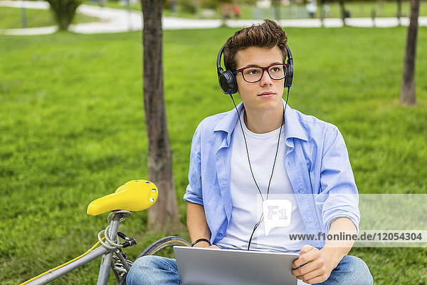 Portrait of serious young man with racing cycle sitting on a bench using laptop and headphones
