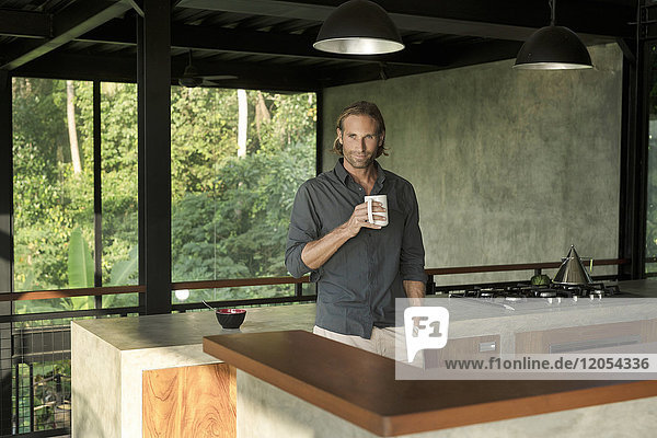 Schöner Mann trinkt Kaffee in moderner Design-Küche mit Glasfassade  umgeben von einem üppigen tropischen Garten.