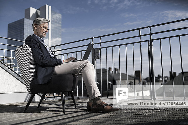 Grauhaariger Geschäftsmann auf dem Balkon sitzend mit Laptop
