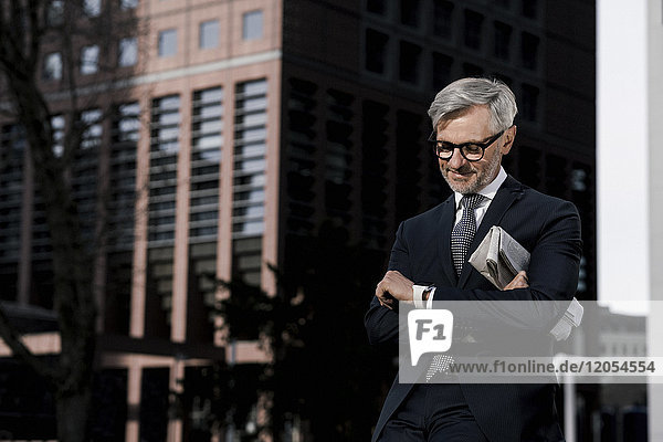 Grauhaariger Geschäftsmann vor rotem Wolkenkratzer mit Blick auf seine smartwatch