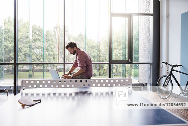 Mann mit Laptop im Pausenraum des modernen Büros auf dem Tischtennistisch