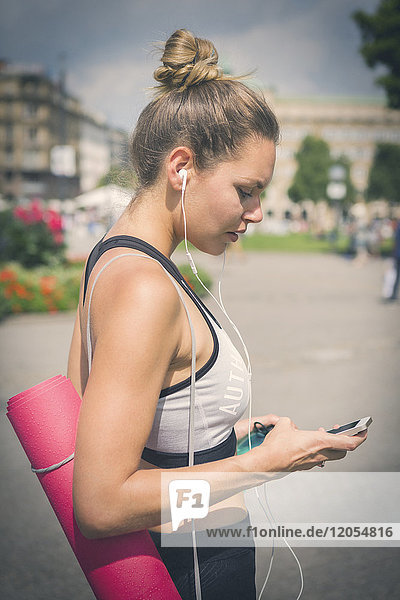 Junge Frau mit Yogamatte  Handy und Kopfhörer in der Stadt