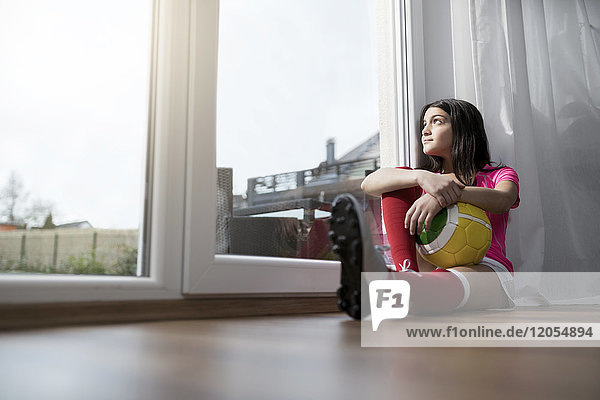 Mädchen im Fußball-Outfit sitzend auf dem Boden im Wohnzimmer mit Blick aufs Fenster