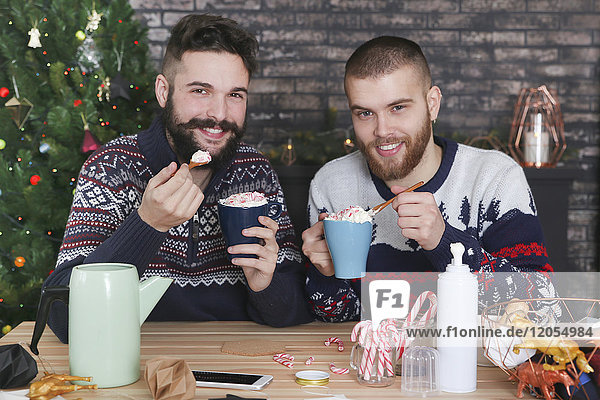 Glückliches schwules Paar trinkt heiße Schokolade mit Sahne und gehackten Zuckerstangen zur Weihnachtszeit.