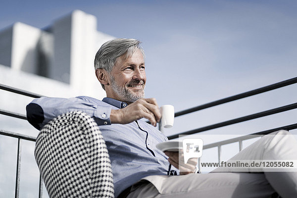 Lächelnder grauhaariger Mann sitzt auf dem Balkon und trinkt Kaffee.