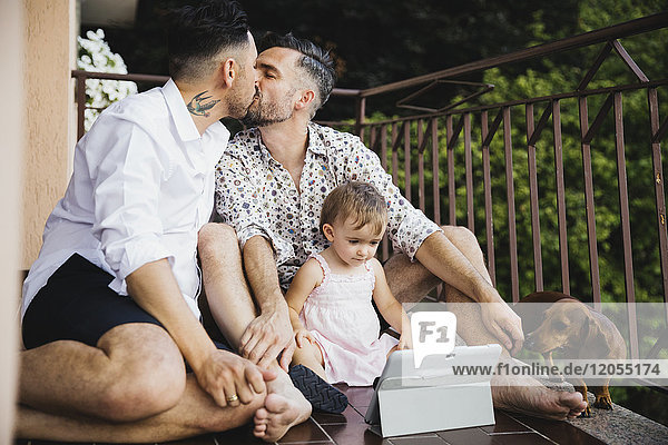 Schwules Paar küssend mit Tochter und Hund auf dem Balkon