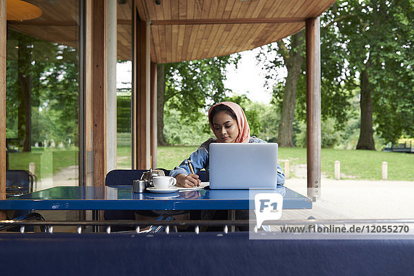 Junge Frau  die Hijab trägt und mit ihrem Laptop auf der Terrasse eines Cafés studiert.