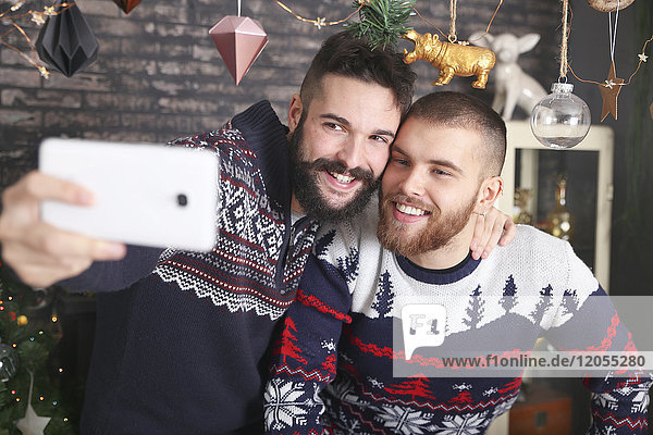 Porträt eines fröhlichen schwulen Paares  das zu Hause mit dem Smartphone unterwegs ist.