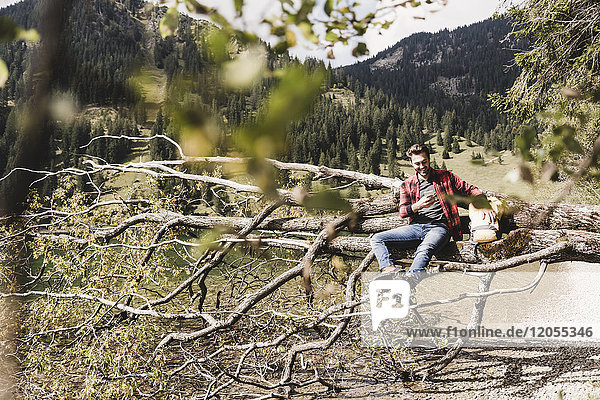 Österreich,  Tirol,  Alpen,  Wanderer entspannt auf Baumstamm am Bergsee beim Handy checken