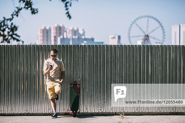 Junger Mann mit Handy an einer Wand gelehnt neben Longboard mit Stadt im Hintergrund