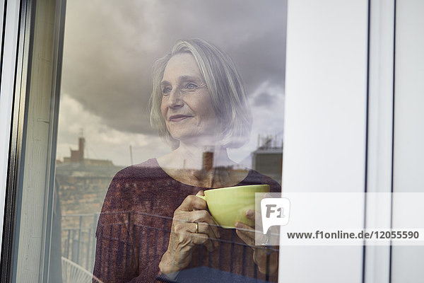 Reife Frau mit Kaffeetasse  die aus dem Fenster schaut.