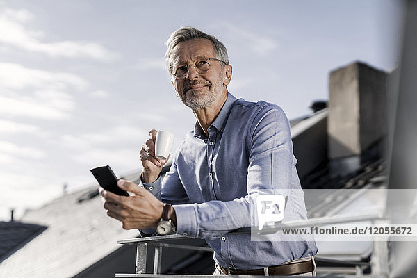 Grey-haired businessman with smartphone standing on balkony drinking coffee