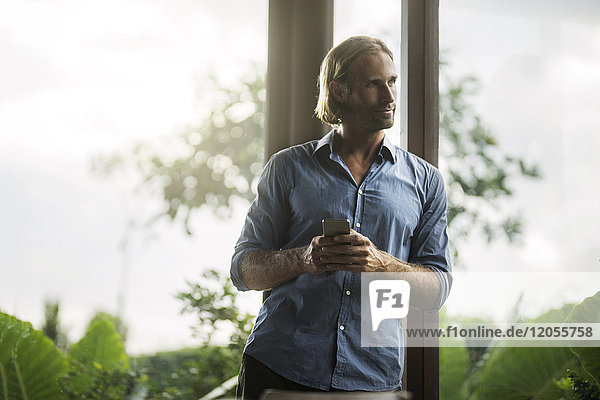 Hübscher Mann mit Smartphone in einem modernen Designhaus mit Glasfassade  umgeben von einem üppigen tropischen Garten.