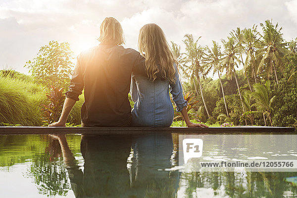 Umarmendes Paar  das am Rande eines Pools sitzt und einen atemberaubenden Blick auf den Sonnenuntergang in einem üppigen tropischen Garten genießt.
