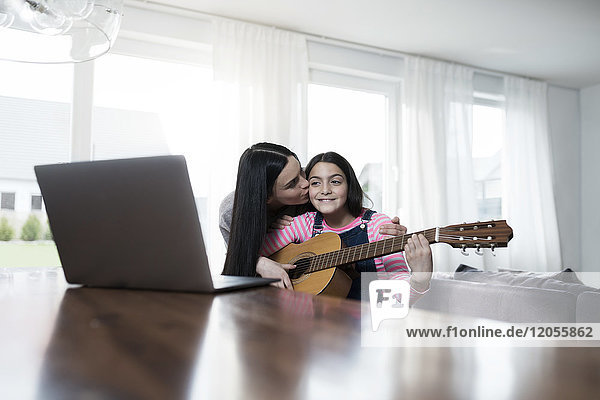 Mother kissing daugher playing guitar in front of laptop
