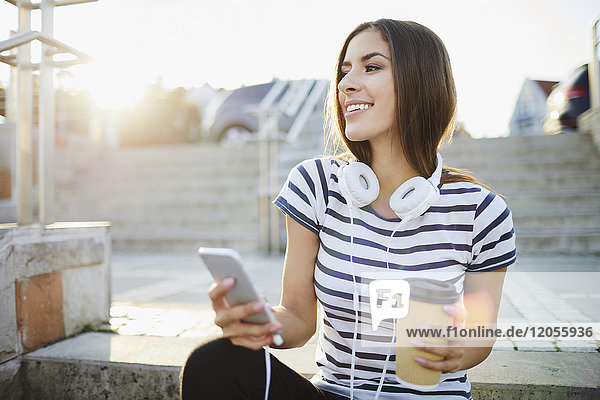 Frau mit Kopfhörer sitzt auf der Treppe in der Stadt mit Smartphone und Kaffee