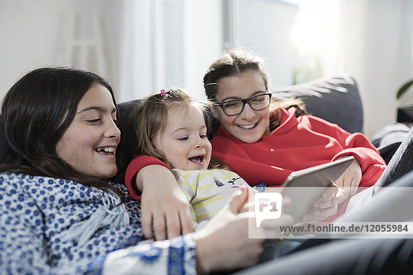 Drei Schwestern lächeln und halten Tablette auf Sofa im Wohnzimmer
