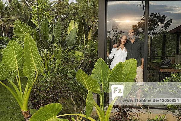 Gartenansicht eines lächelnden Paares  das außerhalb seines Design-Hauses inmitten eines üppigen tropischen Gartens steht.