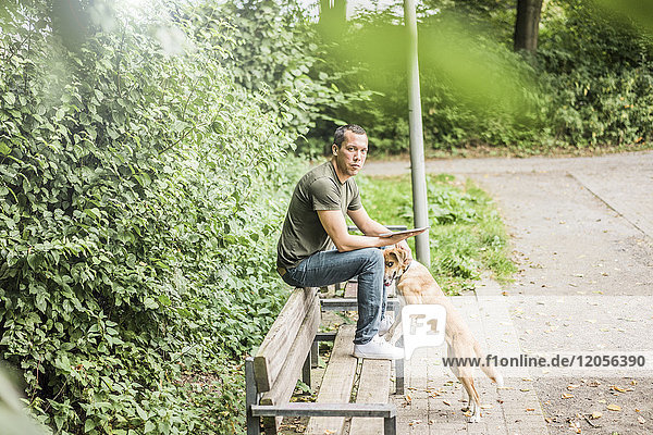 Porträt eines ernsthaften Mannes mit Tablette und Hund auf einer Bank im Park