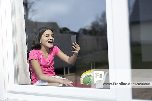 Fröhliches Mädchen im Fußball-Outfit sitzt auf dem Boden im Wohnzimmer und nimmt einen Selfie