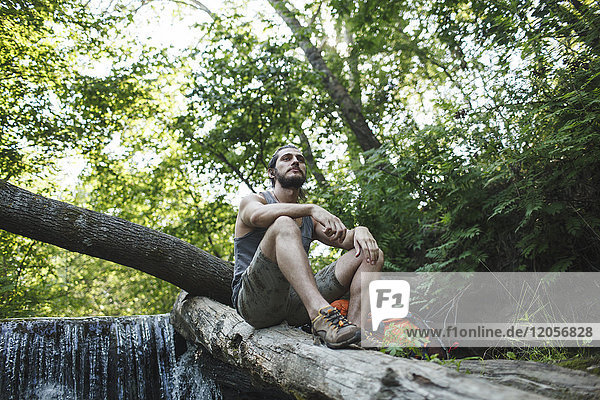 Junger Mann sitzt auf einem Baumstamm an einem Wasserfall im Wald